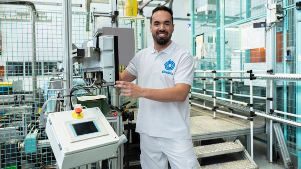 Una mujer con uniforme blanco de Balay conversa con dos visitantes en una luminosa instalación industrial llena de equipos y maquinaria.