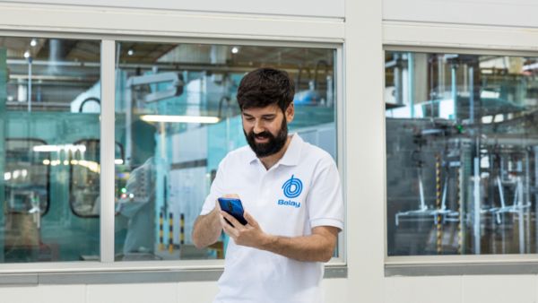 Dos hombres vestidos de manera informal están de pie en una cocina, uno de ellos sosteniendo un teléfono inteligente. 