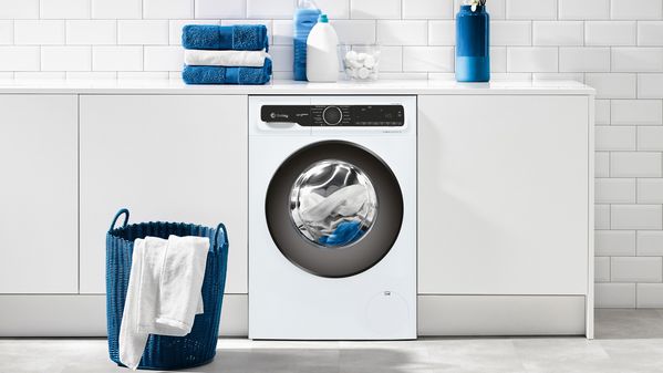 Una lavadora blanca en una lavandería moderna, rodeada de toallas azules dobladas, botellas de detergente y una canasta de ropa azul.