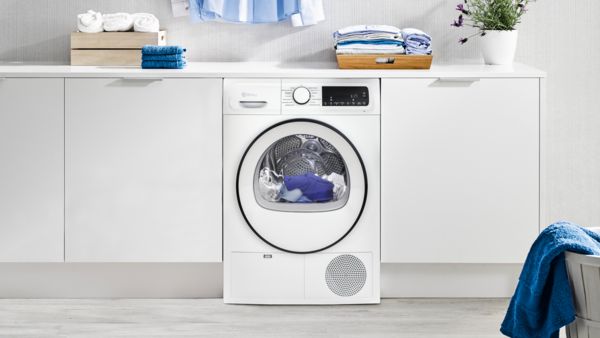 Una secadora blanca en una lavandería moderna, rodeada de toallas azules dobladas, botellas de detergente y una canasta de ropa azul.
