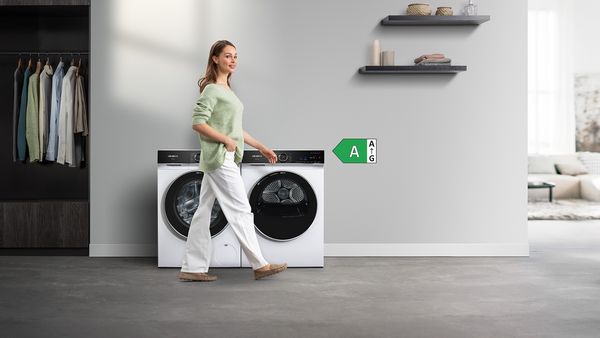 Eine iQ700 Waschmaschine und ein Trockner stehen vor einer grauen Wand. Eine Dame in weißer Hose und grünem Pullover geht daran vorbei.