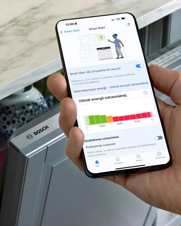Ręka trzymająca smartfon pokazujący cykl mycia naczyń w aplikacji Home Connect od Bosch. W tle widoczna zmywarka.