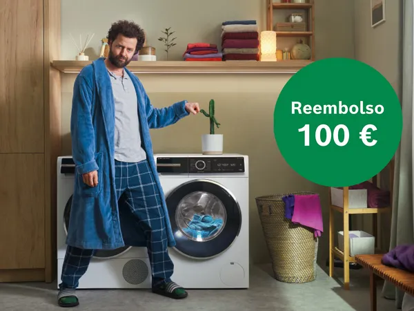 Hombre en bata y pijama señala una planta en una lavadora moderna, rodeado de toallas y una cesta de ropa. Un círculo verde muestra un reembolso de 100 €.