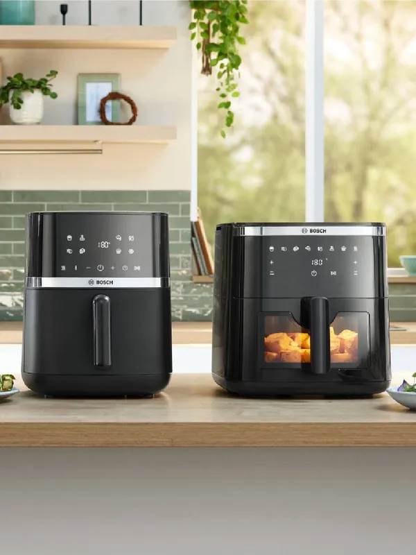 Due friggitrici ad aria Bosch nere sono posizionate sul piano di lavoro in legno della cucina e presentano design eleganti e pannelli di controllo digitali, con luce naturale che filtra.