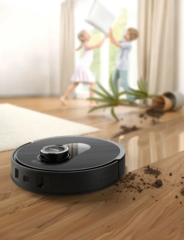 Un robot aspirapolvere su pavimenti in legno, vicino a bambini che giocano e a una pianta in vaso, con lo sporco visibile sul pavimento lì vicino.