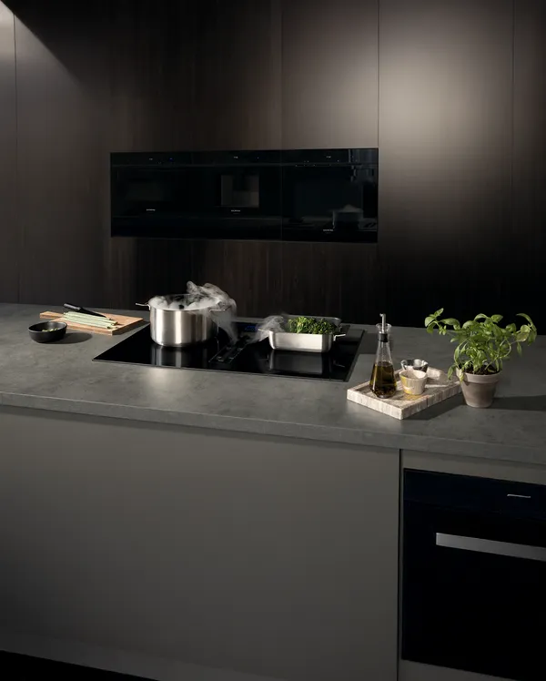 A modern kitchen countertop with a pot steaming, a stainless steel pan with greens, olive oil, and a potted plant on display.