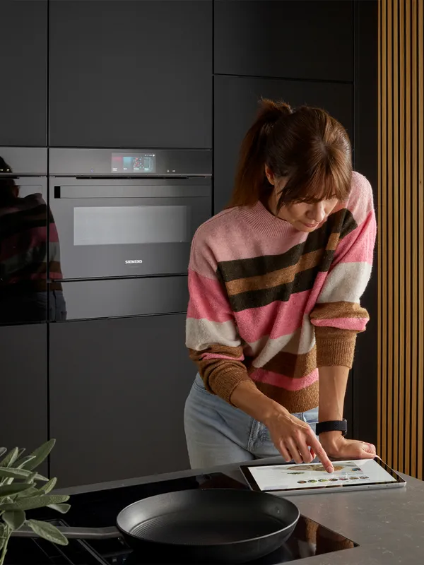 A woman in a pink striped sweater uses a tablet to view a recipe in a modern kitchen with a Siemens oven and induction cooktop.