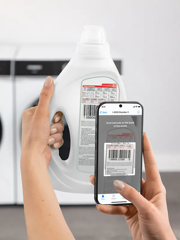A person holds a laundry detergent bottle while scanning its barcode with a smartphone app in a laundry room setting.
