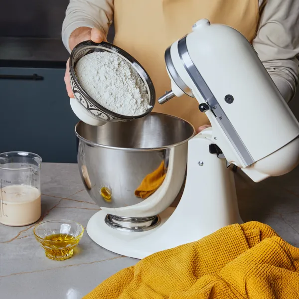 Eine Person gibt Mehl aus einer Schüssel in die Rührschüssel einer Küchenmaschine.
