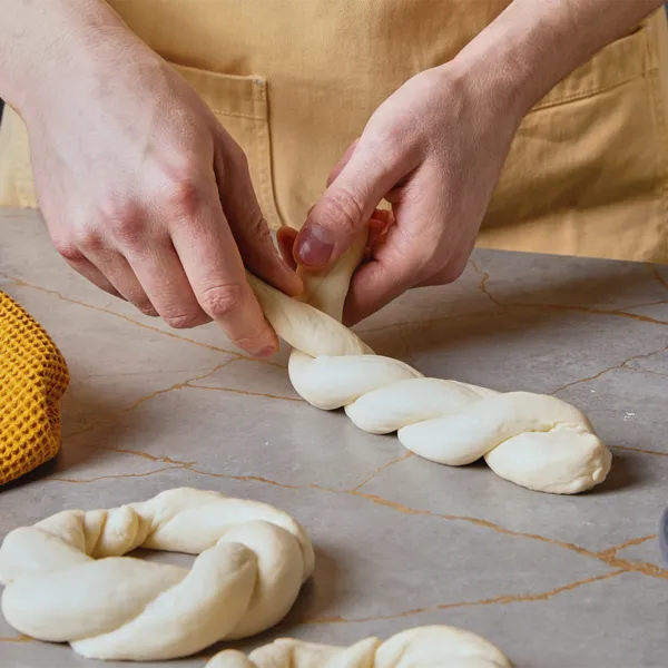 Zwei Hände verflechten zwei Teigstränge auf einer Küchenarbeitsplatte.