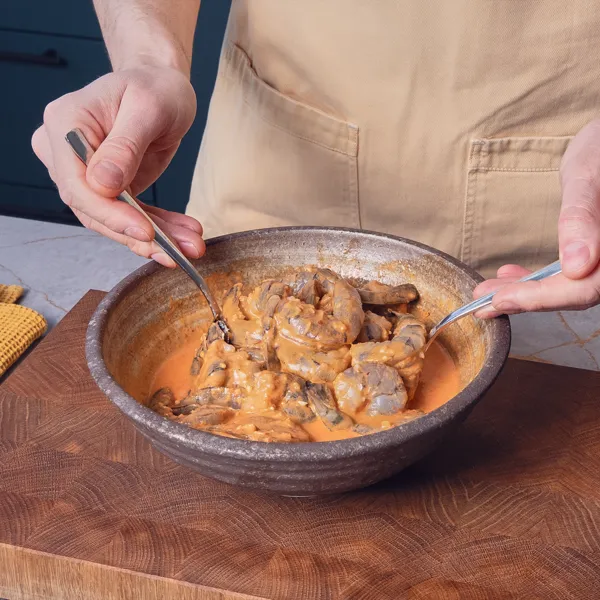 Due mani mescolano i gamberetti e una salsa marrone in una ciotola appoggiata su un tagliere.