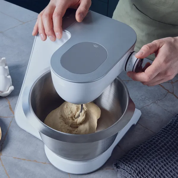 Eine Person verwendet eine Küchenmaschine, um Teig zu kneten, daneben Eier und Butter auf der Küchenarbeitsplatte.