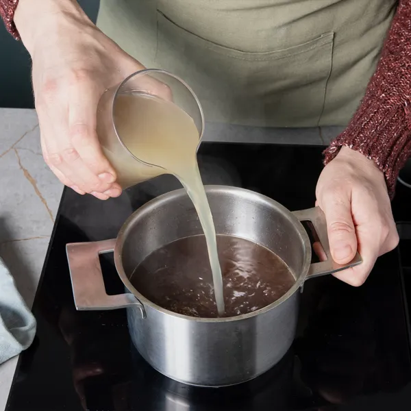 Sauce simmert in einem Kochtopf auf dem Kochfeld, Hühnerbrühe wird unter ständigem Rühren hinzugegeben.
