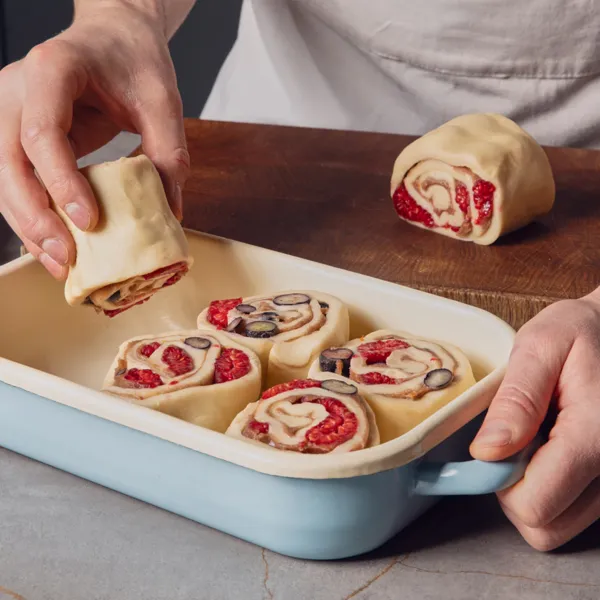 Eine Hand legt ungebackene Zimtbrötchen, aus denen Beeren hervorblitzen, in eine rechteckige blaue Backform auf einer Küchenarbeitsplatte.
