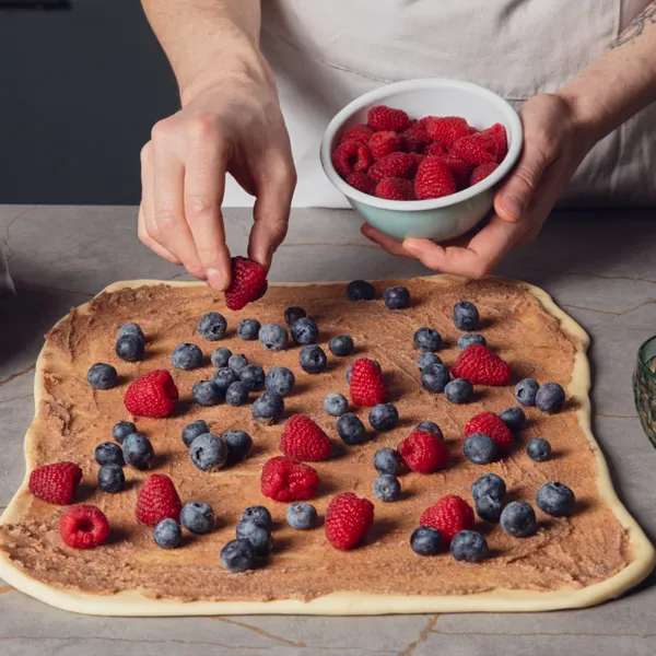 Eine Hand legt Himbeeren auf einen ausgerollten Teig mit brauner Creme auf einer Küchenarbeitsplatte.