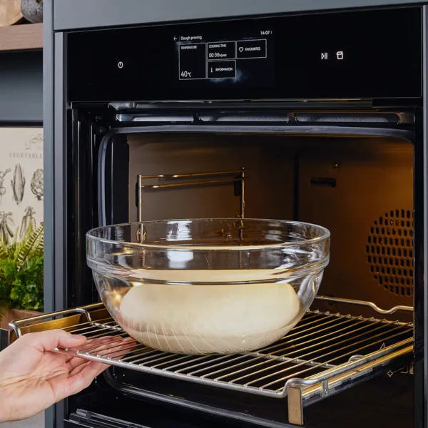 Una mano posiziona una ciotola di vetro con impasto in un forno moderno con display digitale, sullo sfondo di una cucina elegante.