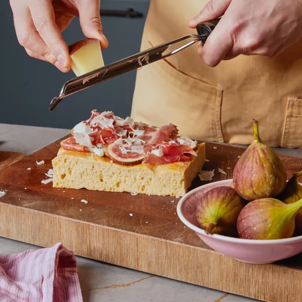 Due mani grattugiano formaggio su un pezzo di focaccia che si trova su un tagliere di legno.