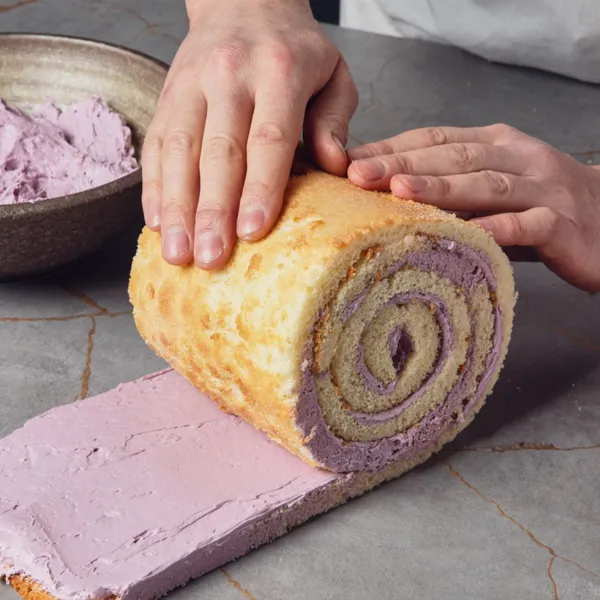 Sul ripiano di una cucina, due mani arrotolano una striscia di impasto cotto ricoperto di crema lilla.