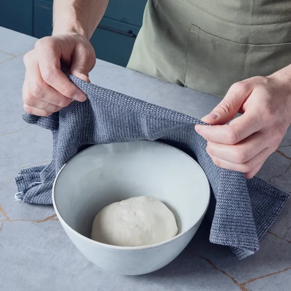 Eine Person deckt eine Schüssel mit Teig auf einer Küchenarbeitsplatte mit einem Geschirrtuch ab, damit der Teig aufgehen kann.
