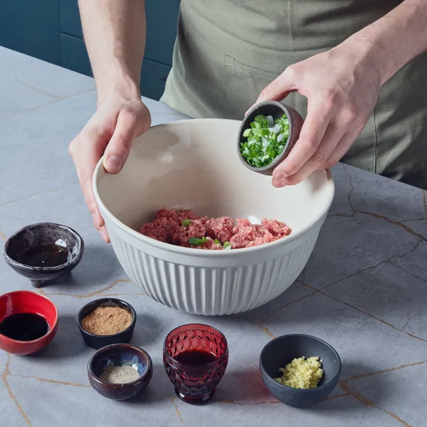 Eine Person gibt zerkleinerte Frühlingszwiebeln in eine Schüssel mit Hackfleisch, daneben kleine Schüsseln mit Saucen und gemahlenem Ingwer für die Schweinefleischfüllung der Teigtaschen.