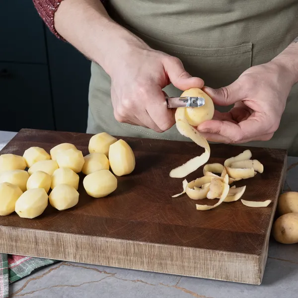 Eine Person schält Kartoffeln auf einer Küchenarbeitsplatte, daneben geschälte Kartoffeln auf einem Holzschneidebrett und eine weihnachtliche Tischdecke.