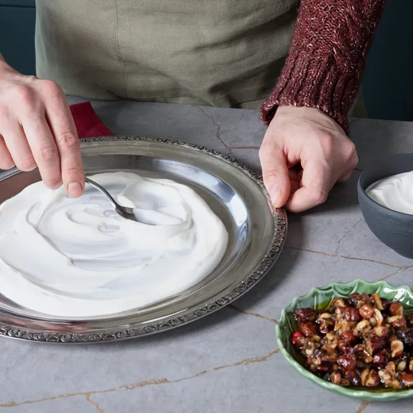 Joghurt wird auf einer silberfarbenen Servierplatte verteilt, daneben eine Schüssel mit Haselnüssen und eine rote Serviette.