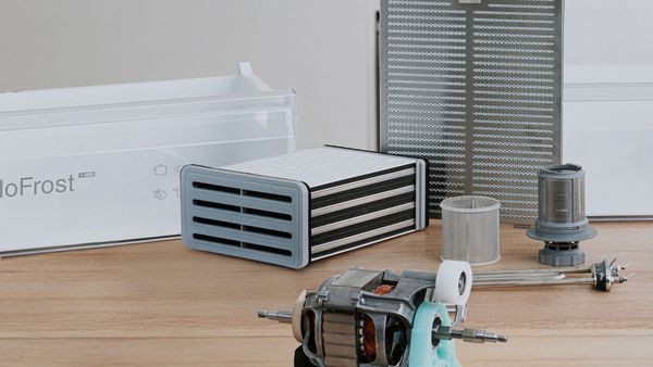 Varios repuestos de electrodom&eacute;sticos, como un filtro de campana o un balc&oacute;n de frigor&iacute;fico, sobre una encimera de madera.