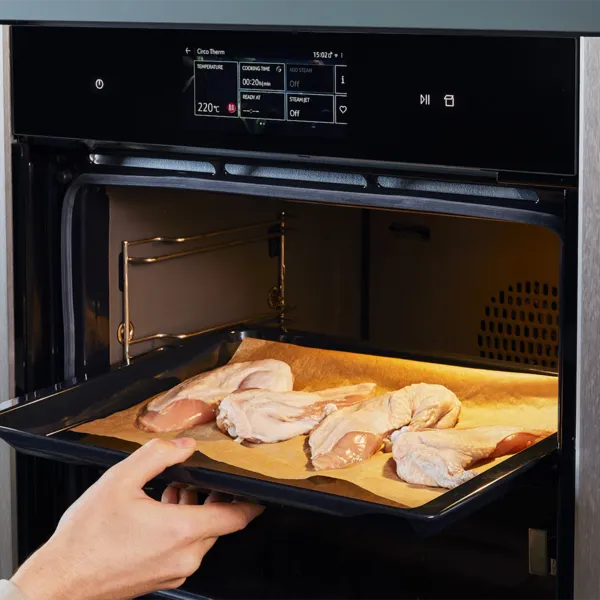 Una mano che fa scivolare una teglia rivestita di carta forno con pezzi di pollo crudo in un forno da incasso Neff; un vaso di fiori e decorazioni da cucina visibili sul bancone.