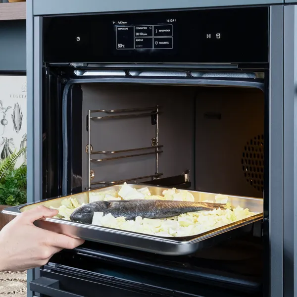 Eine Person schiebt ein Blech mit Wolfsbarsch auf zerkleinertem Kohl zum Garen in einen Dampfbackofen.