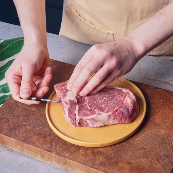 Eine Hand steckt ein kleines Fleischthermometer in die Seite eines rohen Steaks, das auf einem Holzschneidebrett liegt.