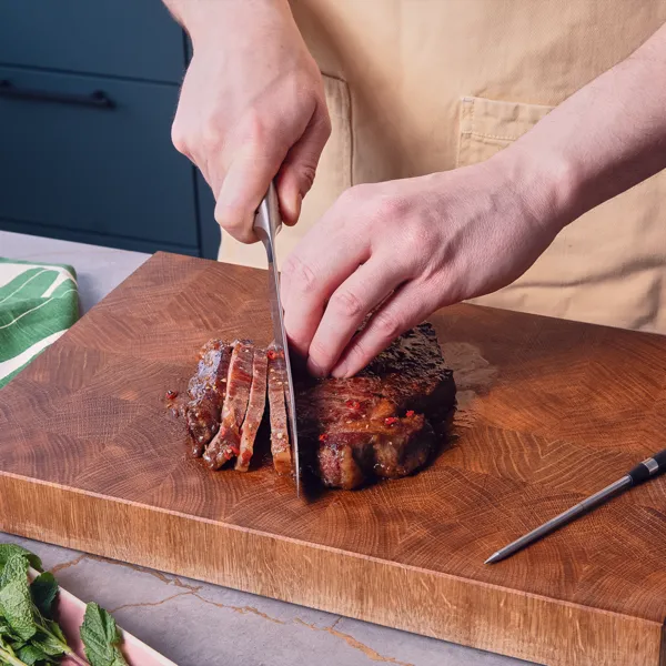 Zwei Hände schneiden ein gegrilltes Steak auf einem Holzschneidebrett in Streifen.
