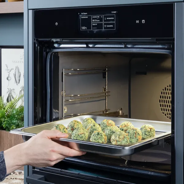 Eine Person schiebt ein Blech mit vorbereiteten Spinat- und Käse-Knödel zum Garen in einen Dampfbackofen.