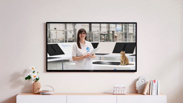 Imagen de televisi&oacute;n colgada en pared sobre mueble bajo de sal&oacute;n y decoraci&oacute;n, mostrando a una empleada de Balay en zona ficticia de pruebas mirando sonriente a c&aacute;mara junto a una encimera ** con una placa mate y un gato naranja.