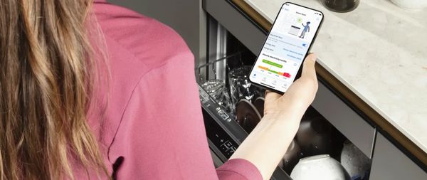 Eine Person h&auml;lt ein Smartphone, auf dem eine App f&uuml;r smarte Geschirrsp&uuml;ler angezeigt wird, w&auml;hrend sie neben einer offenen, mit Geschirr gef&uuml;llten Sp&uuml;lmaschine steht. 