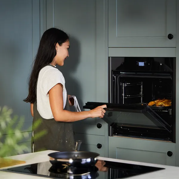 A person in a kitchen opens an oven door, revealing freshly baked goods, with modern cabinetry and a stovetop in the foreground.