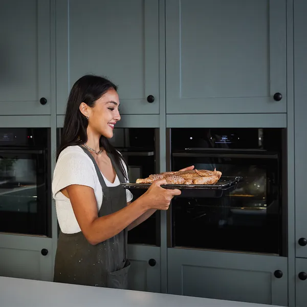 Woman in apron removing baked goods from oven in modern kitchen.