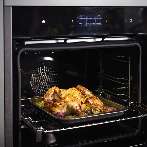 A roasted chicken with herbs and vegetables is placed on a tray inside a modern oven, displaying a digital control panel.