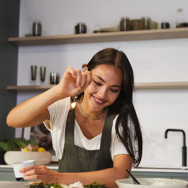 Smiling woman in a green apron adds seasoning to a dish with a spoon in a kitchen.
