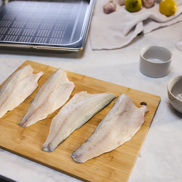 Wooden cutting board with raw fish fillets, baking tray, and various cooking ingredients on a kitchen counter.