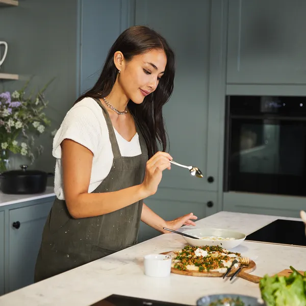 Woman in a green apron tasting food with a spoon in a modern kitchen with green cabinets and built-in ovens.
