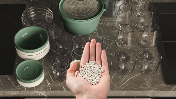 Hand met zeolieten korrels boven een marmeren aanrecht met glazen en servies