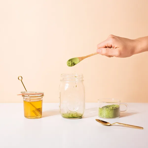 Hand sprinkling green matcha powder from a wooden spoon into a glass jar, with honey jar and cup of green powder on white surface.