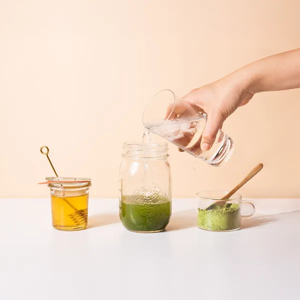 Hand pouring water into a jar with green matcha powder, next to a jar of honey with a dipper and a cup of matcha powder with a wooden spoon.