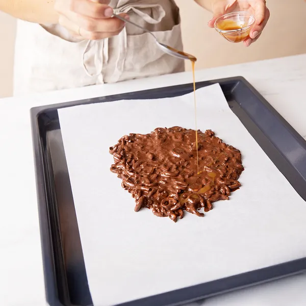 Pouring melted chocolate over crushed pretzels and caramel on a baking sheet to make chocolate pretzel bark