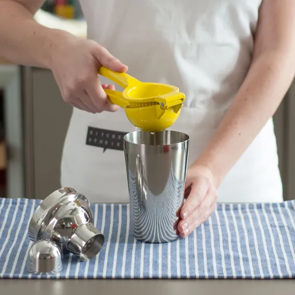 Person squeezing a yellow citrus juicer over a metal cocktail shaker on a striped cloth.
