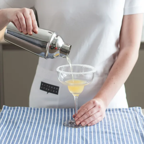 A person in a white apron pours a yellow drink from a silver cocktail shaker into a sugar-rimmed glass