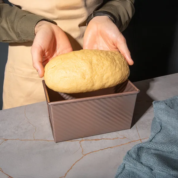 Hands holding a large, golden-brown loaf of bread placed in a rectangular baking pan on a marble surface.