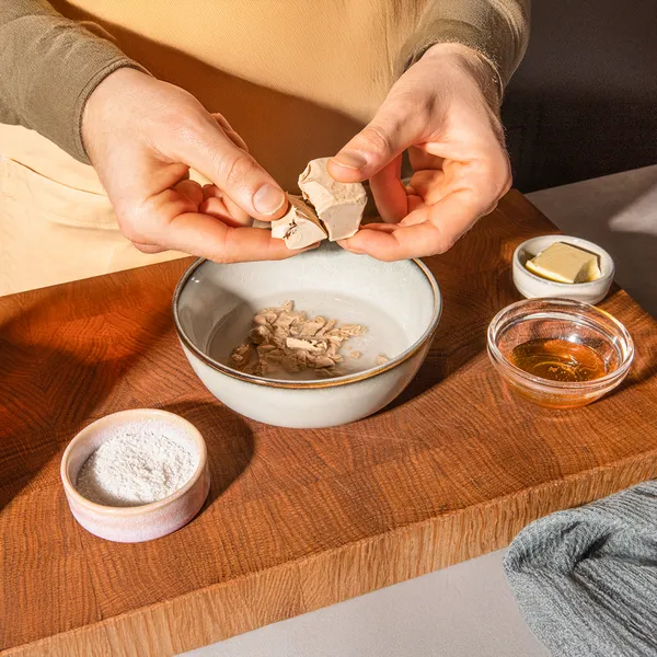 Hands preparing food ingredients on a wooden board, including a bowl, powder, and other cooking elements.