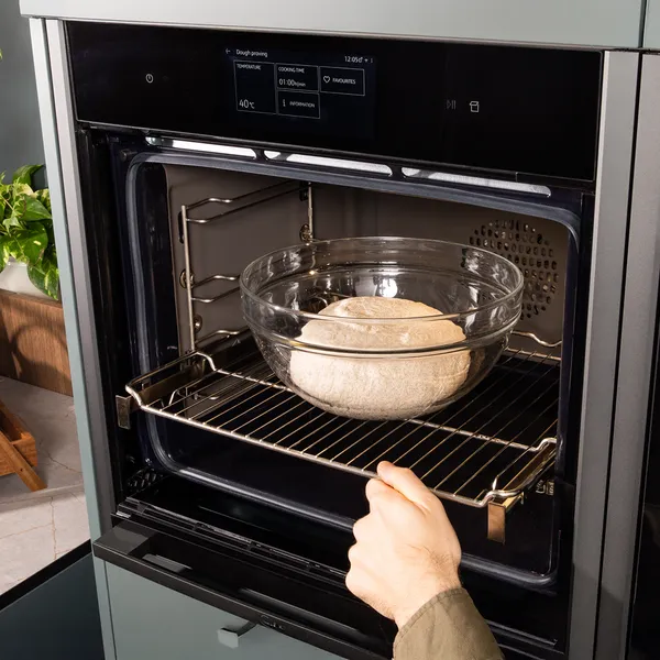 Oven with baking dish inside, hand reaching to remove dish, kitchen counter with plants and utensils.