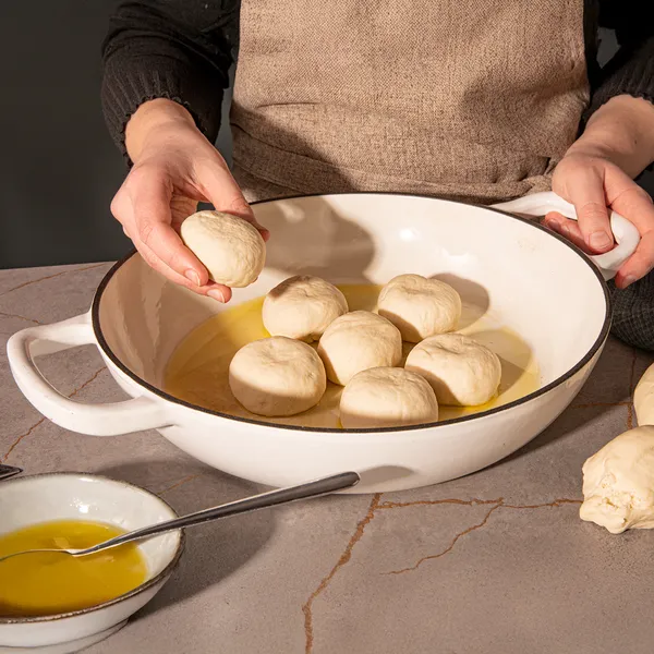Hands preparing homemade dumplings in a bowl with oil on a wooden table.